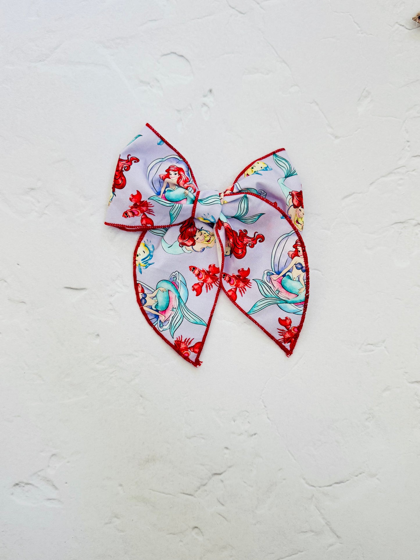 Midi-sized bow featuring an Ariel-inspired mermaid print with sea characters, red serged edges, and an alligator clip.